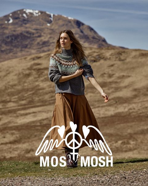 Eine Frau in einem gemusterten Pullover und einem braunen Rock steht im Freien, hinter ihr sind Hügel und ein Berg zu sehen. Am unteren Bildrand ist das „MOSS MOSH“-Logo zu sehen.