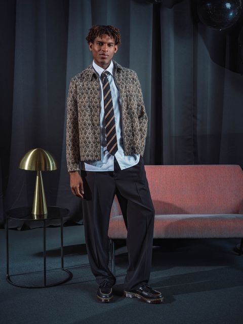 Ein Mann in einer gemusterten Jacke, Krawatte und Anzughose steht neben einem rosa Sofa und einer goldenen Lampe vor dunklen Vorhängen.