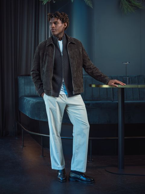 Ein Mann in einer braunen Jacke und einer weißen Hose steht neben einem runden Tisch in einer modernen, schwach beleuchteten Lounge mit dunkelblauen Sitzmöbeln und Vorhängen.