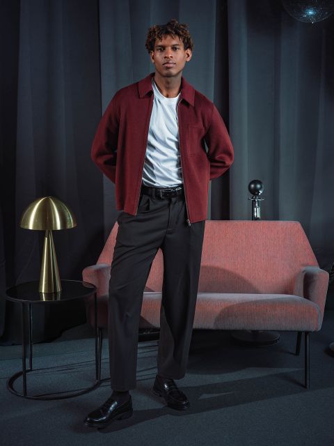 Ein Mann in einer burgunderroten Jacke und einer schwarzen Hose steht neben einem rosa Sofa, auf dem ein runder Tisch mit einer goldenen Lampe steht, vor dunklen Vorhängen.
