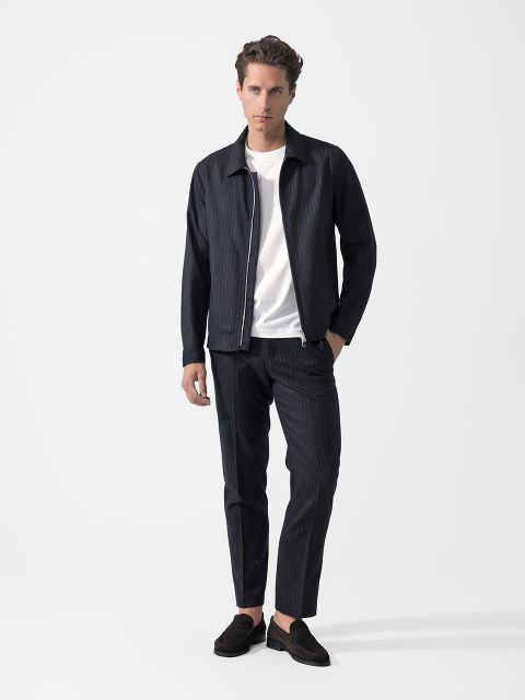Vor einem weißen Hintergrund steht ein Mann, der eine dunkle Nadelstreifenjacke, eine dazu passende Hose, ein weißes T-Shirt und schwarze Slipper trägt.