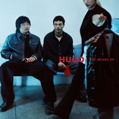 Zwei Männer sitzen auf einer Bank, während eine Person im Vordergrund eine Kleidersack mit der Aufschrift „HUGO RED MEANS GO“ in der Hand hält.