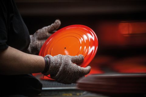 Eine Person, die Handschuhe trägt, hält einen leuchtend orangefarbenen, runden Metalldeckel in den Händen; im Hintergrund sind verschwommene orangefarbene Gegenstände zu sehen.
