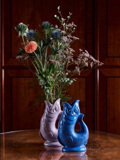 Zwei fischförmige Vasen, eine violette mit Blumen und eine blaue, stehen auf einem Holztisch vor einem Hintergrund aus dunklem Holzvertäfelung.