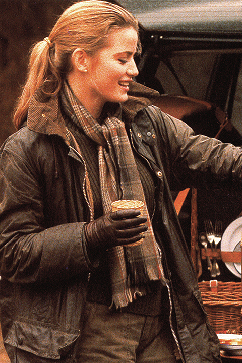 Eine lächelnde Frau in Jacke, Schal und Handschuhen hält im Freien neben einem Picknickkorb und Besteck eine Tasse in der Hand; ihr Haar ist zu einem Pferdeschwanz zusammengebunden.