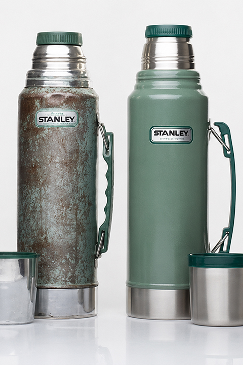 Zwei Stanley-Thermoskannen stehen nebeneinander; die eine ist verwittert und verrostet, die andere ist sauber und neu. Beide haben einen grünen Korpus und einen Metalldeckel.