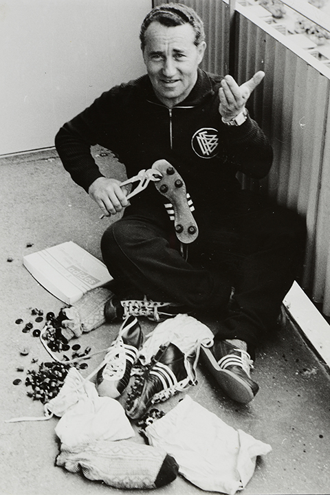 Ein Mann sitzt auf dem Boden und hält einen Fußballschuh in der Hand; um ihn herum liegen Schuhe, Schrauben und Socken; er lächelt und gestikuliert mit der Hand.