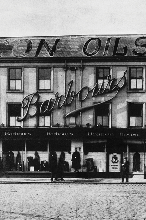Schwarz-Weiß-Foto eines Gebäudes mit einem großen „Barbours“-Schild und darunter liegenden Ladenfronten; davor stehen Menschen, darüber ist ein Teil des „BEACON OILS“-Schilds zu sehen.