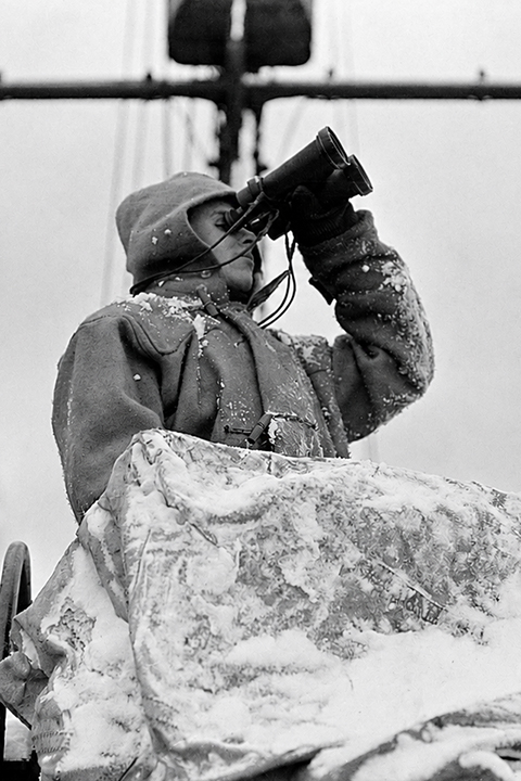Eine Person in dicker Winterkleidung, mit Schnee bedeckt, die im Freien sitzend durch ein Fernglas schaut, im Hintergrund sind Maschinen zu sehen.
