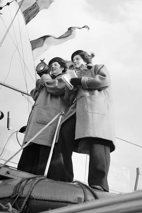 Zwei Frauen in Mänteln stehen auf dem Deck eines Bootes; die eine hält eine Lampe in der Hand, die andere ein Notizbuch, während über ihnen vor dem Hintergrund eines bewölkten Himmels Fahnen wehen.