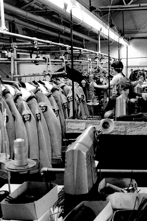Arbeiter in einer Fabrik hängen Jacken an einen Kleiderständer und prüfen sie, umgeben von Nähmaschinen und anderen Geräten.