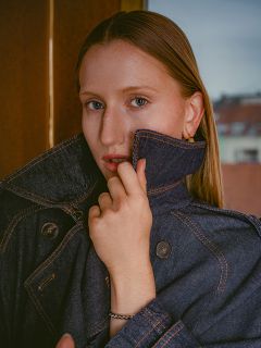 Eine Frau mit glattem Haar trägt eine dunkle Jeansjacke, deren Kragen sie hochgeschlagen hat, und blickt mit ausdruckslosem Gesicht in die Kamera.