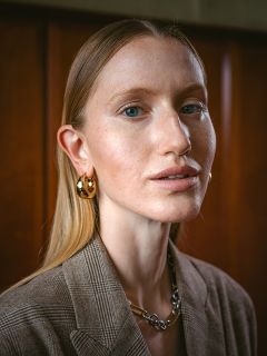 Eine Frau mit glattem blondem Haar, goldenen Creolen und einem braunen karierten Blazer blickt vor einem Holzhintergrund in die Kamera.