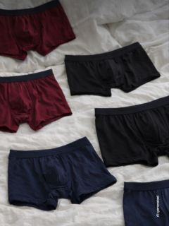 Sechs Paar Boxershorts für Herren in den Farben Schwarz, Burgunderrot und Marineblau liegen auf einer weißen Stoffunterlage ausgebreitet.