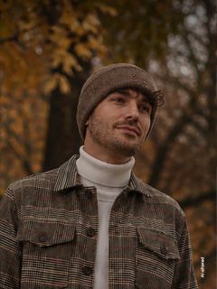 Ein Mann mit einer braunen Mütze und einer karierten Jacke steht im Freien vor dem Hintergrund herbstlicher Bäume, blickt zur Seite und lächelt leicht.