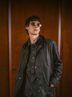 Eine Person mit Sonnenbrille und dunkler Jacke steht vor Holztüren, schaut leicht zur Seite und hat die Hände in den Taschen.