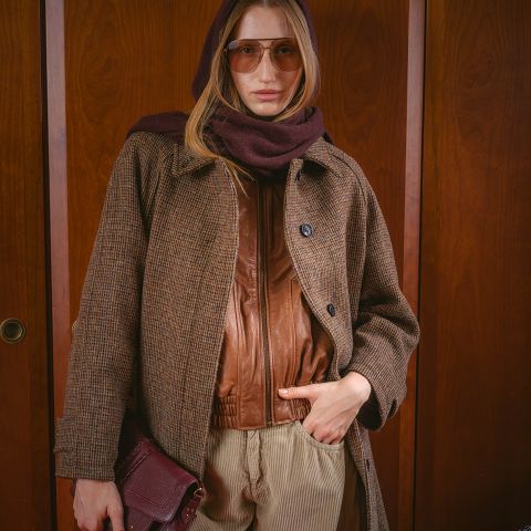 Eine Frau mit Sonnenbrille, einem braunen Schal, einem Mantel und einer Lederjacke, die eine Handtasche in der Hand hält und vor einem hölzernen Hintergrund steht.