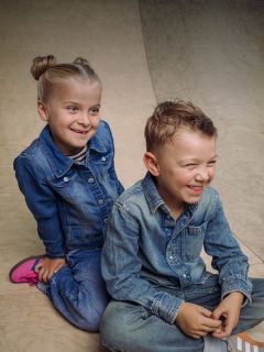 Zwei kleine Kinder in Jeansoutfits sitzen auf einer Holzbank, lächeln und lachen miteinander. Das Mädchen hat blonde Haare, die zu Zöpfen geflochten sind; der Junge hat kurze Haare.