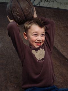 Ein lächelnder kleiner Junge in einem braunen Sweatshirt hält einen Basketball über den Kopf und steht auf einer Skate-Rampe, im Hintergrund sind Graffiti zu sehen.
