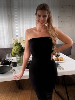 Eine Frau in einem trägerlosen schwarzen Kleid steht lächelnd neben einem Esstisch, der mit Blumen, Weintrauben, Gebäck und Wein gedeckt ist, in einem modernen, hell erleuchteten Raum.
