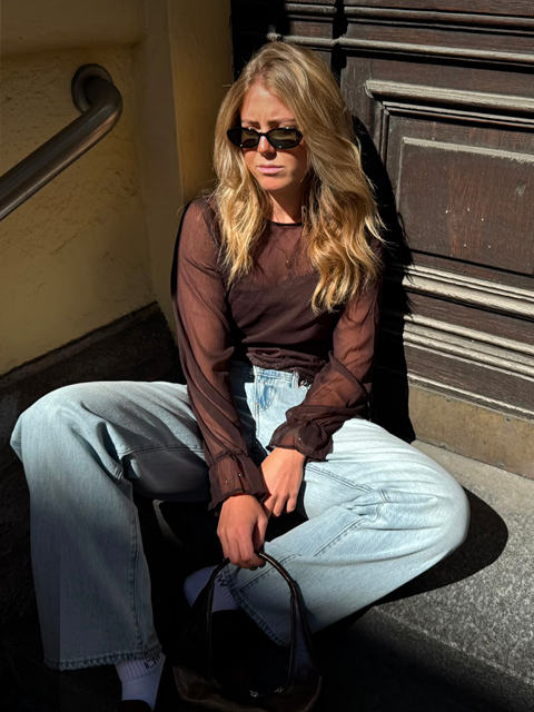 Eine Frau mit Sonnenbrille, einem durchsichtigen schwarzen Oberteil und hellblauen Jeans sitzt auf einer sonnigen Steinstufe neben einer dunklen Holztür und hält eine schwarze Tasche in der Hand.
