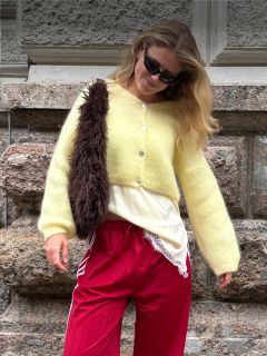 Eine Frau mit schwarzer Sonnenbrille, gelber Strickjacke, roter Hose und einer braunen, pelzbesetzten Umhängetasche steht vor einer strukturierten Steinmauer und lächelt leicht.