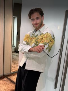Ein bärtiger Mann trägt eine helle Jacke mit gelben Blumenmustern, hält eine Sonnenbrille in der Hand und steht in einem modernen Raum mit einem zotteligen Teppich.