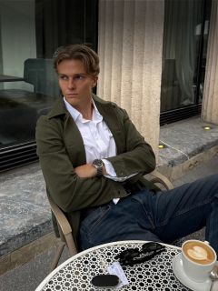 Ein Mann in einem weißen Hemd und einer grünen Jacke sitzt an einem Tisch im Straßencafé, vor sich stehen Kaffee, eine Sonnenbrille und Schlüssel, und er schaut zur Seite.