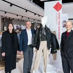 Four adults stand in a clothing store beside a mannequin wearing layered outerwear and a scarf, smiling at the camera.