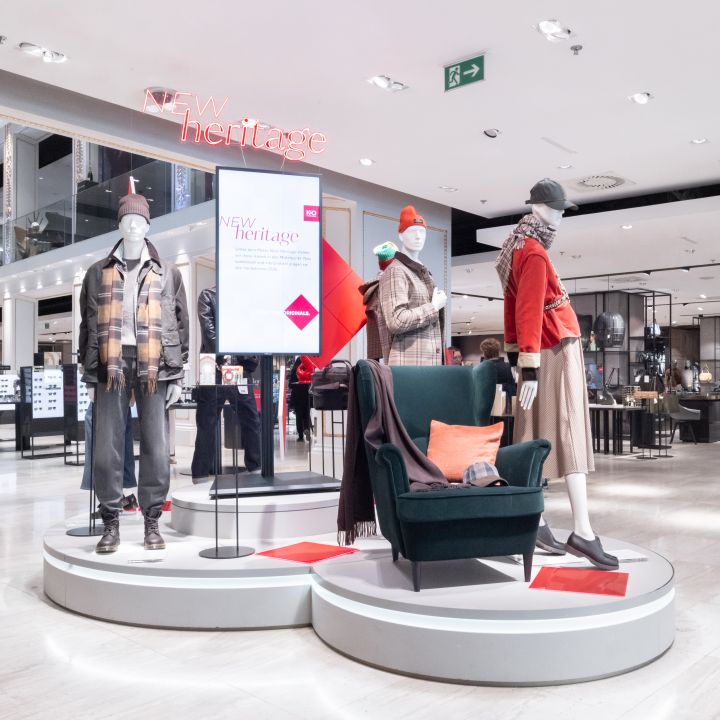Mannequins in winter outfits on a circular white platform in a mall window, with a neon 'NEW heritage' sign overhead.