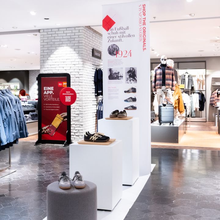 Retail store interior displaying shoes on white pedestals with mannequins in the background and a brick wall feature in a bright, modern space.