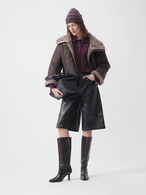 Eine Frau mit Mütze, Lammfelljacke, burgunderrotem Hemd, Leder-Culottes und Stiefeln mit Absatz, die eine marineblaue Tasche in der Hand hält und vor einem weißen Hintergrund steht.