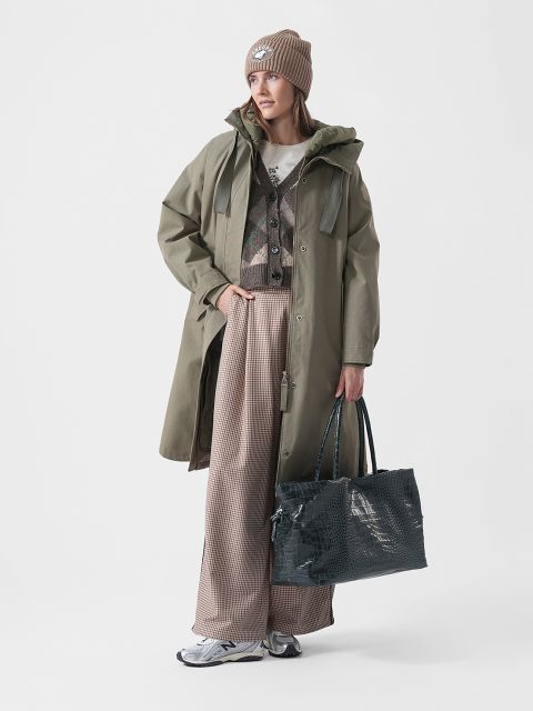 Eine Frau in einem grünen Mantel, gemusterten Hosen, Sneakers und einer Mütze hält eine große, strukturierte Tasche in der Hand und steht vor einem schlichten weißen Hintergrund.