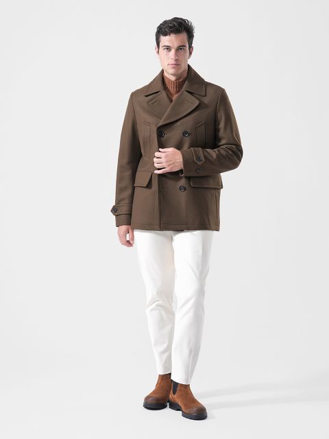 Ein Mann, der einen braunen Mantel, einen hellbraunen Pullover, eine weiße Hose und braune Stiefel trägt, steht vor einem schlichten weißen Hintergrund.