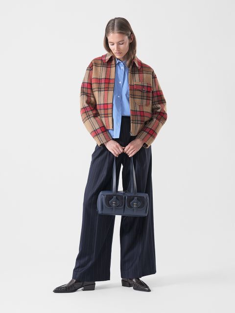 Eine Frau in einer karierten Jacke, einem blauen Hemd und einer Hose mit breiten Nadelstreifen hält eine marineblaue Handtasche mit zwei Fächern in der Hand und steht vor einem schlichten weißen Hintergrund.