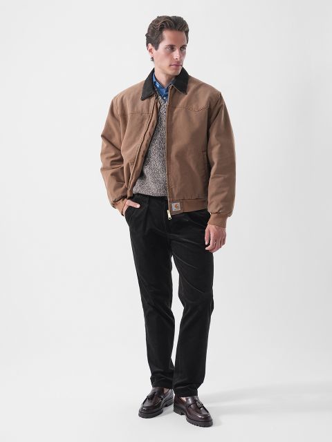 Ein Mann, der eine braune Jacke, einen grauen Pullover, ein blaues Hemd, eine schwarze Hose und dunkle Schuhe trägt und vor einem schlichten weißen Hintergrund steht.