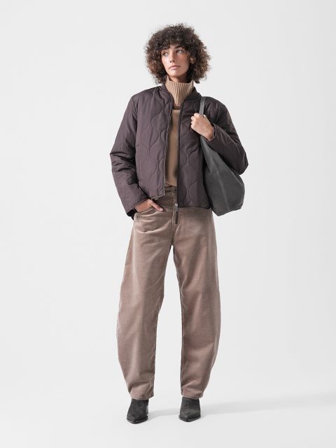 Eine Person in einer braunen Steppjacke, einem hellbraunen Rollkragenpullover, einer weiten beigen Hose und schwarzen Schuhen, die eine schwarze Tragetasche in der Hand hält und vor einem einfarbigen Hintergrund steht.