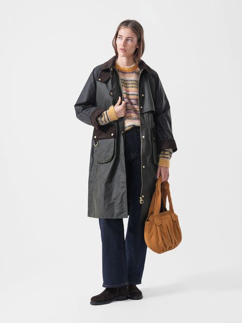 Eine Frau in einem dunklen Mantel, einem gestreiften Pullover, blauen Jeans und braunen Schuhen, die eine senffarbene Handtasche in der Hand hält und vor einem schlichten weißen Hintergrund steht.