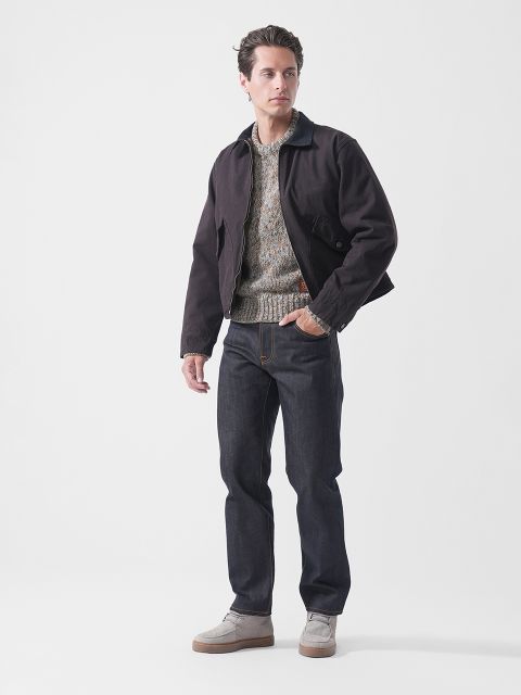 Vor einem schlichten weißen Hintergrund steht ein Mann, der eine dunkle Jacke, einen gemusterten Pullover, dunkle Jeans und helle Schuhe trägt.