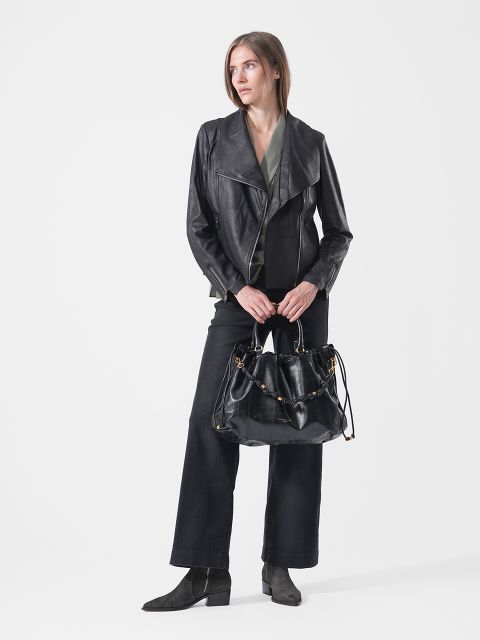 Eine Frau in einer schwarzen Lederjacke, einer schwarzen Hose und Stiefeln steht mit einer großen schwarzen Handtasche in der Hand vor einem weißen Hintergrund und blickt zur Seite.