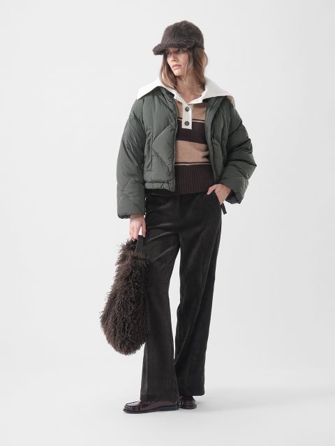 Eine Frau in einer grünen Daunenjacke, einem gestreiften Pullover, einer braunen Hose und einer Pelzmütze, die eine flauschige braune Tasche in der Hand hält und vor einem schlichten weißen Hintergrund steht.