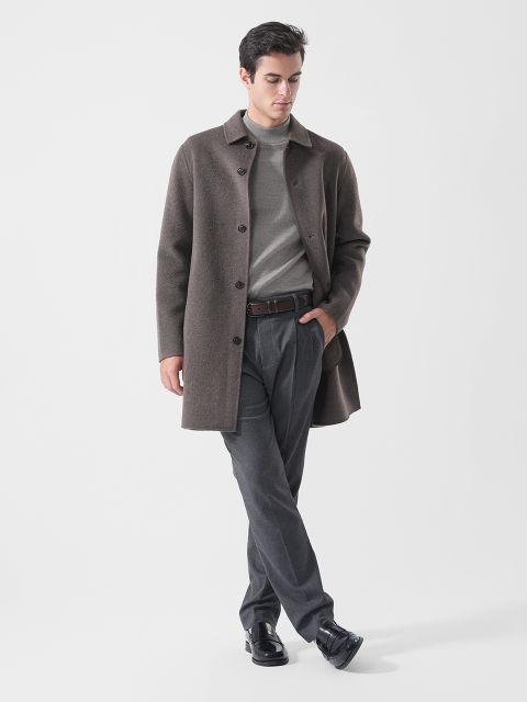 Ein Mann, der einen braunen Mantel, einen grauen Pullover, eine graue Hose und schwarze Schuhe trägt, steht mit entspannten Armen und gekreuzten Beinen vor einem schlichten weißen Hintergrund.