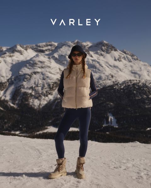 Eine Frau, die eine hellbraune Fleeceweste, marineblaue Leggings, einen Bucket-Hat und Winterstiefel trägt, steht im Schnee vor einer Bergkette unter dem Varley-Logo.