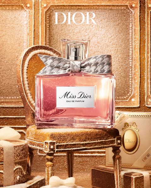Ein Flakon „Miss Dior Eau de Parfum“ mit einer gemusterten Schleife, der in der Mitte eines kleinen, verzierten Hockers steht, der von Geschenkboxen umgeben ist.