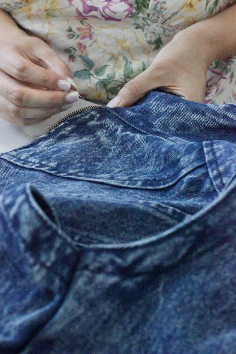 Eine Person, die den Reißverschluss einer blauen Jeans festhält, ein geblümtes Hemd mit gelben und rosa Blumen trägt und deren Hände sowie ein Teil der Jeans zu sehen sind.