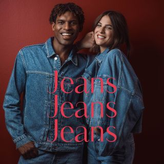 Zwei Personen in blauen Jeansjacken und Jeans stehen lächelnd vor einer roten Wand. Darüber steht in großen rosa Buchstaben der Schriftzug „Jeans Jeans Jeans“.