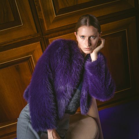Eine Person in einem violetten Kunstpelzmantel lehnt an einem Stuhl, blickt in die Kamera, im Hintergrund sind holzvertäfelte Wände zu sehen.