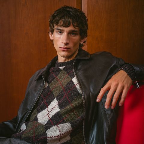 Ein junger Mann in einer dunklen Lederjacke und einem Argyle-Pullover sitzt auf einem roten Stuhl, posiert vor einer Holzwand und blickt in die Kamera.
