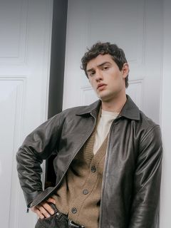 Ein Mann, der eine dunkle Lederjacke über einer hellbraunen Strickjacke und einem cremefarbenen Hemd trägt und an einem Türrahmen lehnt.