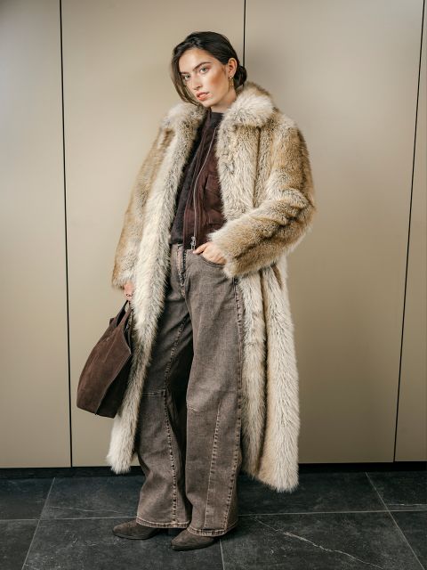 Eine Frau in einem langen Pelzmantel, einer braunen Hose und Stiefeln steht in einem Raum, hält eine braune Handtasche in der Hand und blickt mit ausdruckslosem Gesicht in die Kamera.